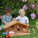 Two children sitting on grass playing with the Ostheimer dolls house placed in front of them. One of the children is holding a Holztiger dinosaur. There are other Holztiger figures in the house and a Bajo tractor in front. These wooden figures are part of a large range of expertly hand crafted, Waldorf inspired, wooden toys here at Babipur.