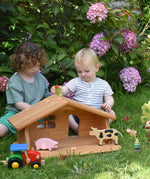 Two children sitting on grass playing with the Ostheimer dolls house placed in front of them. One of the children is holding a Holztiger dinosaur. There are other Holztiger figures in the house and a Bajo tractor in front. These wooden figures are part of a large range of expertly hand crafted, Waldorf inspired, wooden toys here at Babipur.