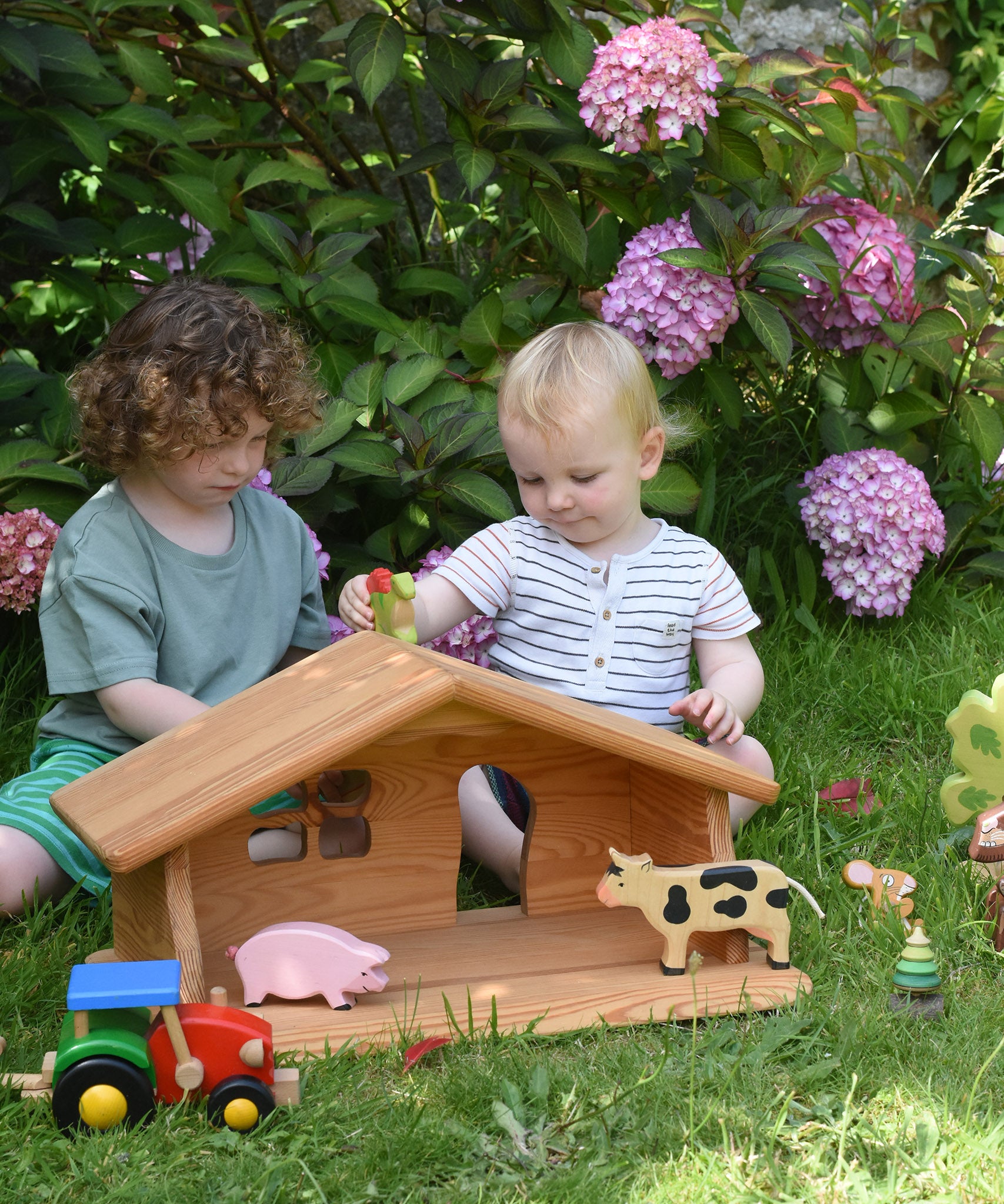 Two children sitting on grass playing with the Ostheimer dolls house placed in front of them. One of the children is holding a Holztiger dinosaur. There are other Holztiger figures in the house and a Bajo tractor in front. These wooden figures are part of a large range of expertly hand crafted, Waldorf inspired, wooden toys here at Babipur.