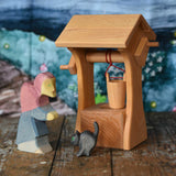 An Ostheimer wooden water well with roof toy placed on a wooden surface with an Ostheimer witch and black car for witch figure. A Wonderie wondercloth can be seen in the background. These wooden figures are part of a wide range of expertly hand crafted wooden toys here at Babipur.  
