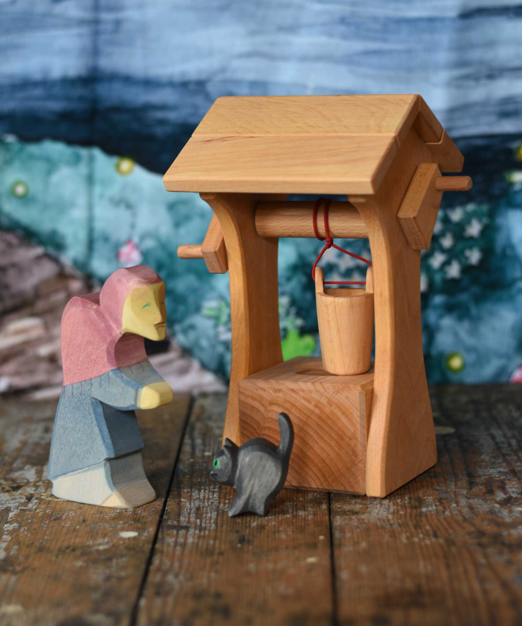 An Ostheimer wooden water well with roof toy placed on a wooden surface with an Ostheimer witch and black car for witch figure. A Wonderie wondercloth can be seen in the background. These wooden figures are part of a wide range of expertly hand crafted wooden toys here at Babipur.  