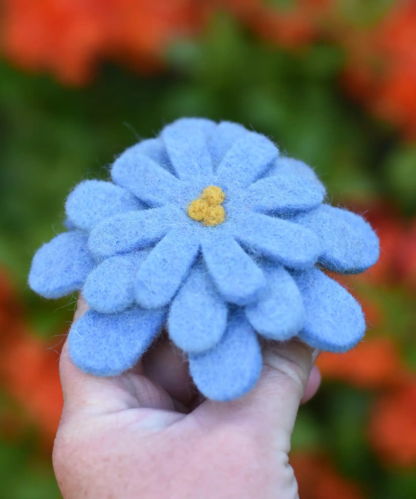 Blue Papoose toys felt aster flower in an adult's hand with orange azalea flowers in the background. 