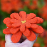 Orange Papoose toys felt aster flower in an adult's hand with orange azalea flowers in the background. 