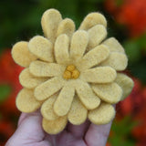 Yellow Papoose toys felt aster flower in an adult's hand with orange azalea flowers in the background. 