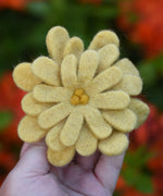 Yellow Papoose toys felt aster flower in an adult's hand with orange azalea flowers in the background. 