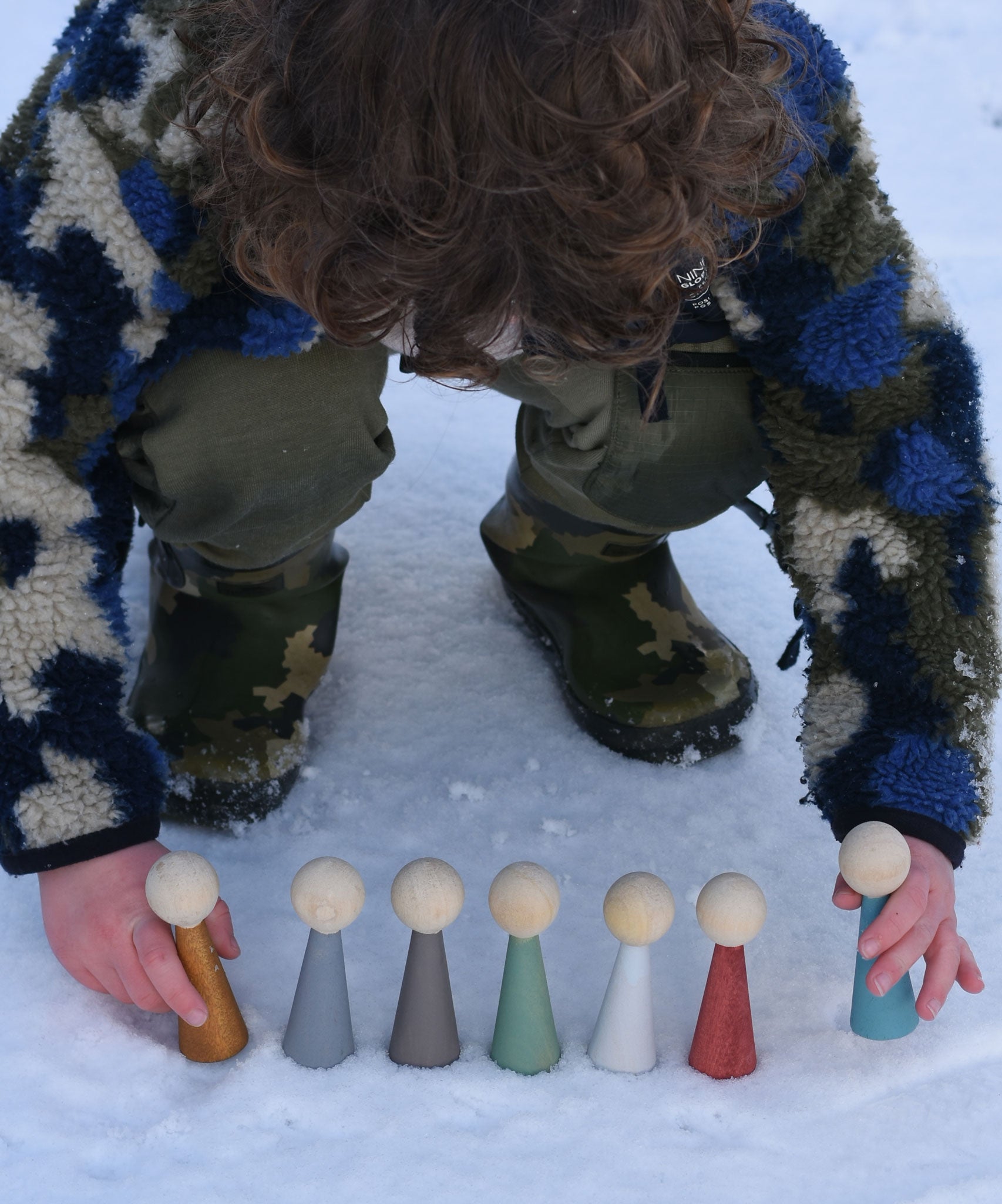 A child playing with the Papoose Toys Wooden Earth colours People peg dolls placed on a snow covered surface outoors.