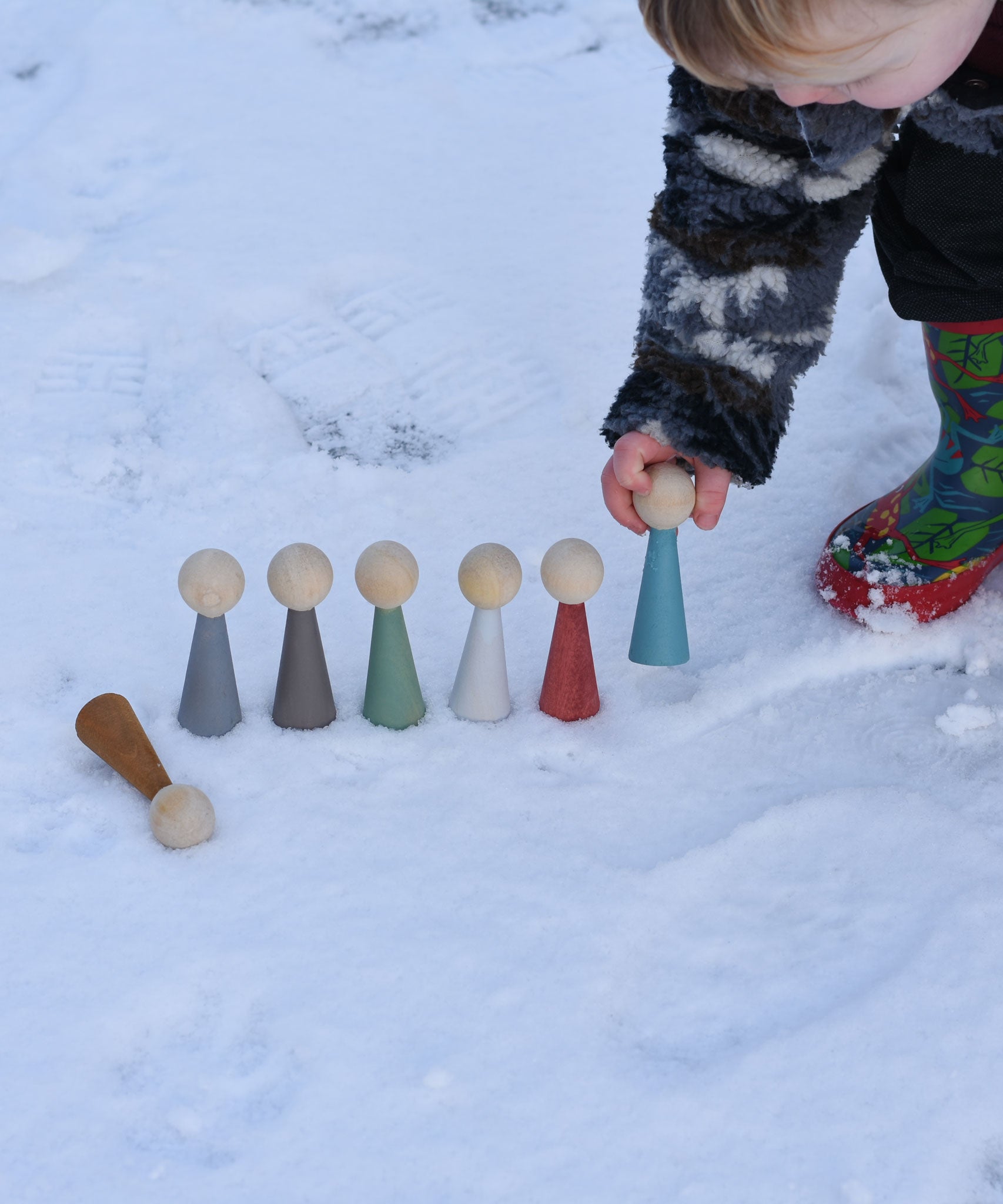 A child playing with the Papoose Toys Wooden Earth colours People peg dolls placed on a snow covered surface outoors.