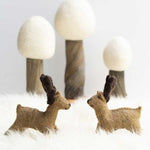 2 Papoose handmade felt reindeer toy figures stood on a white fluffy blanket in front of 3 Papoose winter trees