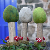 Papoose Toys Summer, Winter and Spring felt woodland trees lined up on a stone wall outdoors. placed on a rattan top stool. 