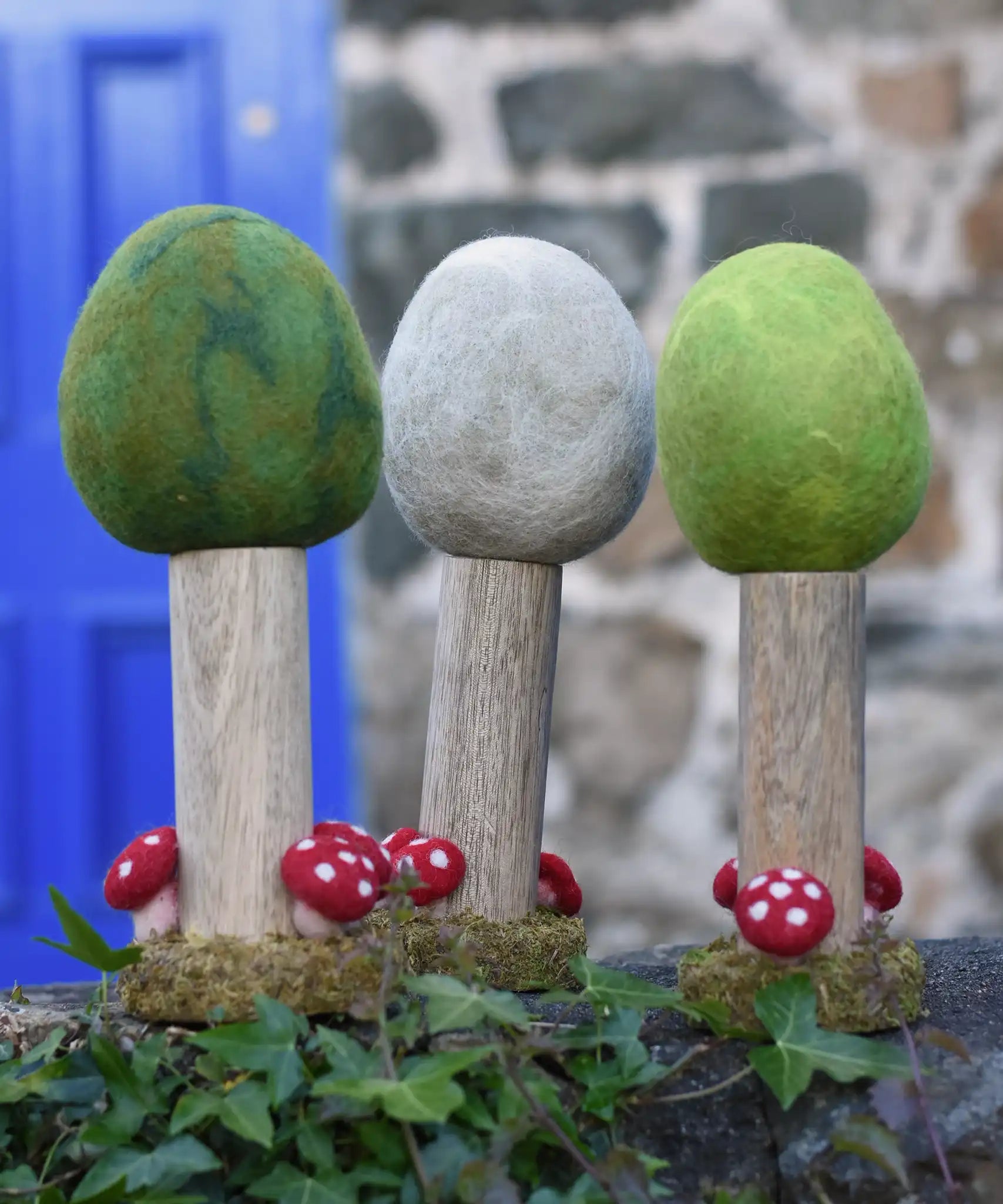 Papoose Toys Summer, Winter and Spring felt woodland trees lined up on a stone wall outdoors. placed on a rattan top stool. 