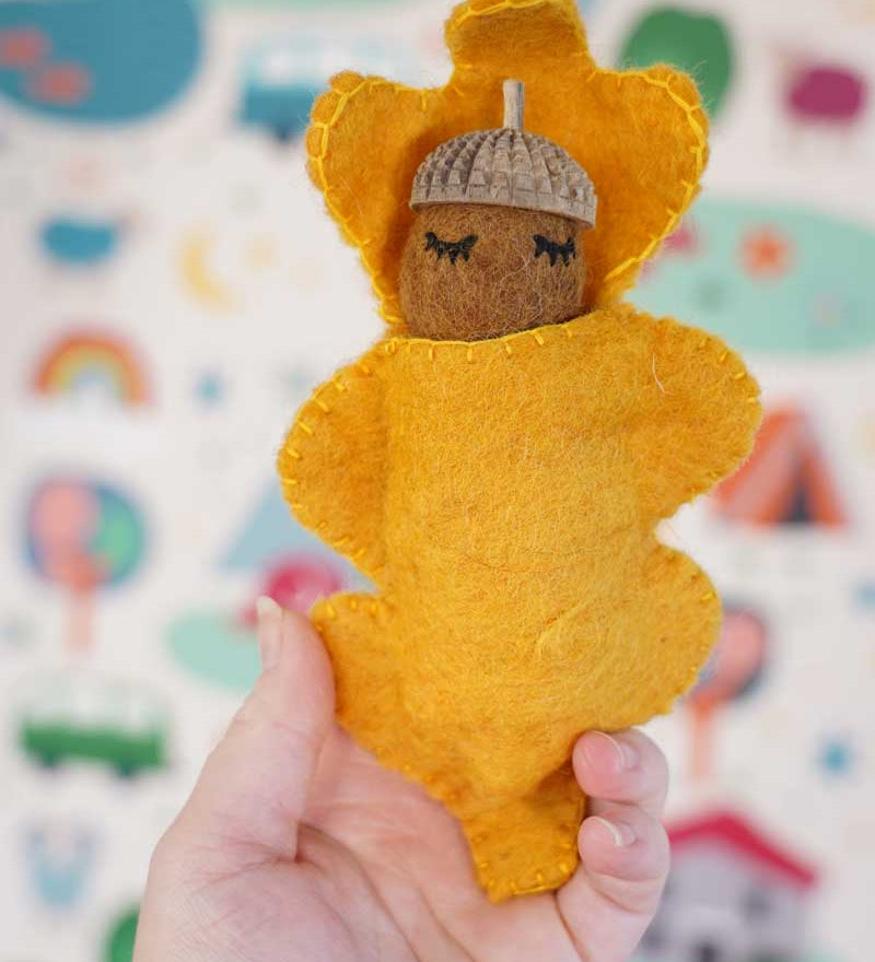 Close up of a hand holding a Papoose handmade felt acorn baby toy figure in front of a patterned wallpaper