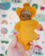 Close up of a hand holding a Papoose handmade felt acorn baby toy figure in front of a patterned wallpaper