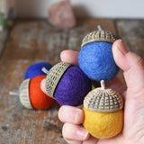 Three Papoose Toys rainbow acorns in an adult's hand, some on a wooden desk in the background. 7 colours of rainbow felt on hand carved wooden acorn caps.