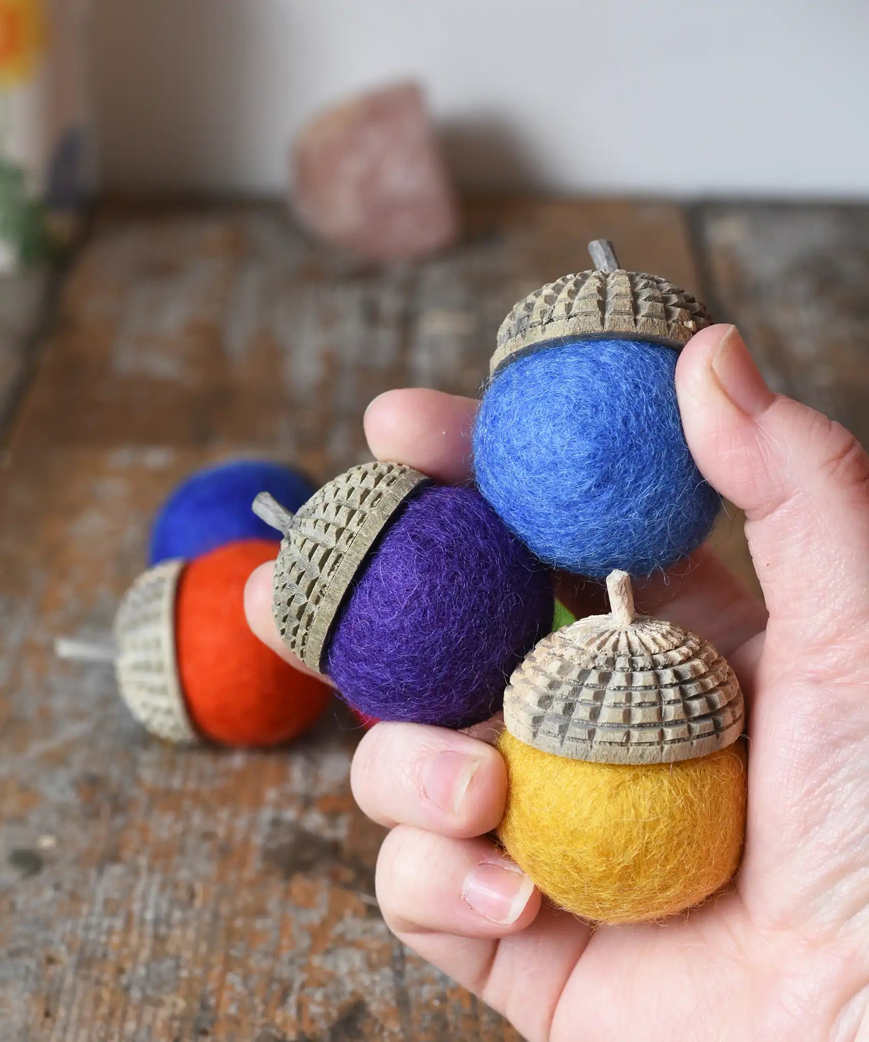 Three Papoose Toys rainbow acorns in an adult's hand, some on a wooden desk in the background. 7 colours of rainbow felt on hand carved wooden acorn caps.