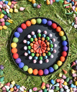 Papoose rainbow felt balls placed in a mandala with Grapat mandala pieces on a black metal tray. 