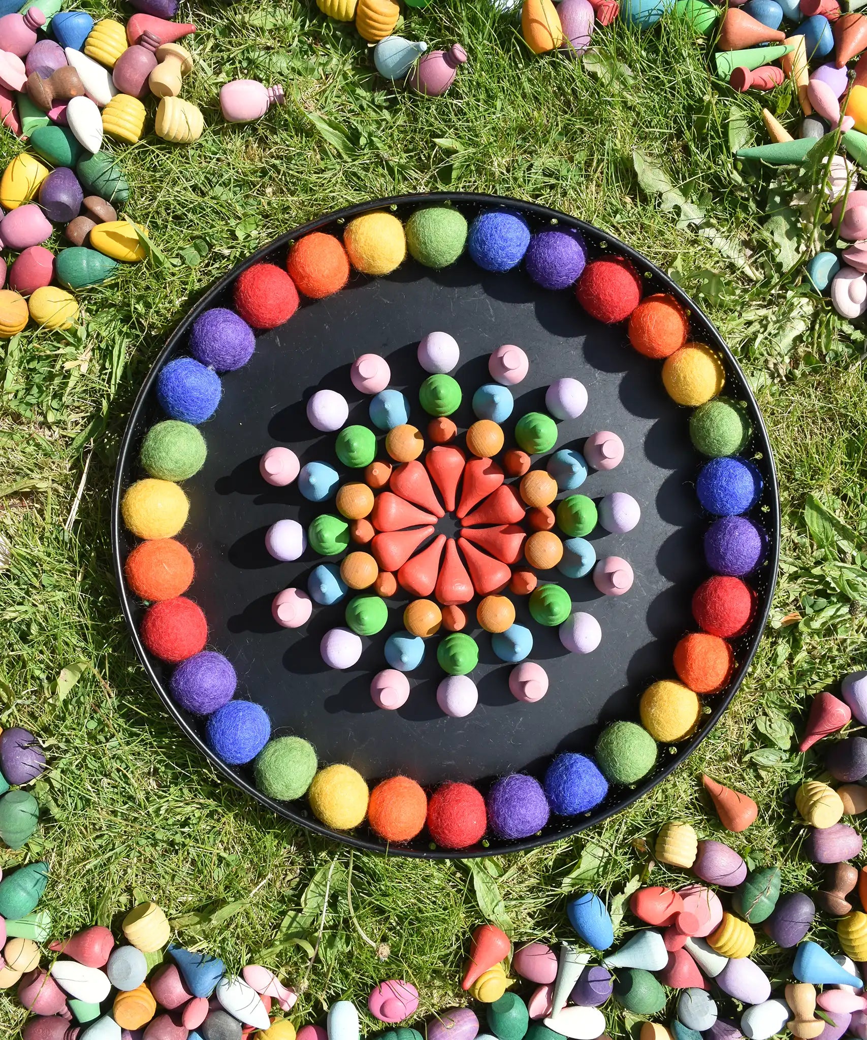 Papoose rainbow felt balls placed in a mandala with Grapat mandala pieces on a black metal tray. 