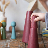 Papoose Toys wooden stacking cones in earth red placed on a wooden desk. Child's hand holding the smallest piece.