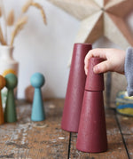 Papoose Toys wooden stacking cones in earth red placed on a wooden desk. Child's hand holding the smallest piece.