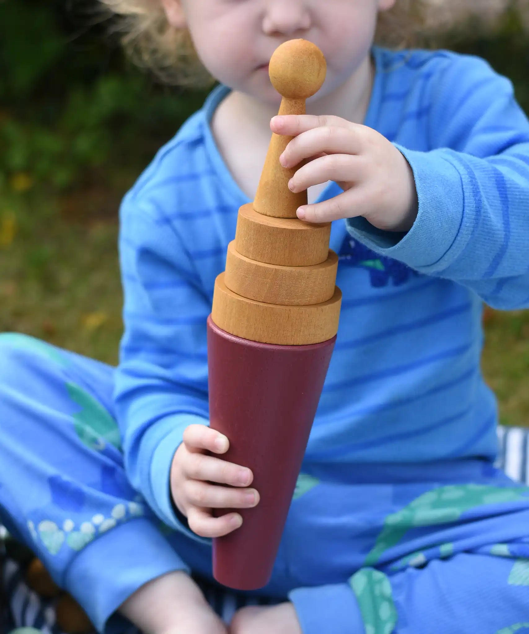 Child using a Papoose Toys wooden stacking cone in earth red as an ice cream cone with yellow discs and a yellow peg doll on top.