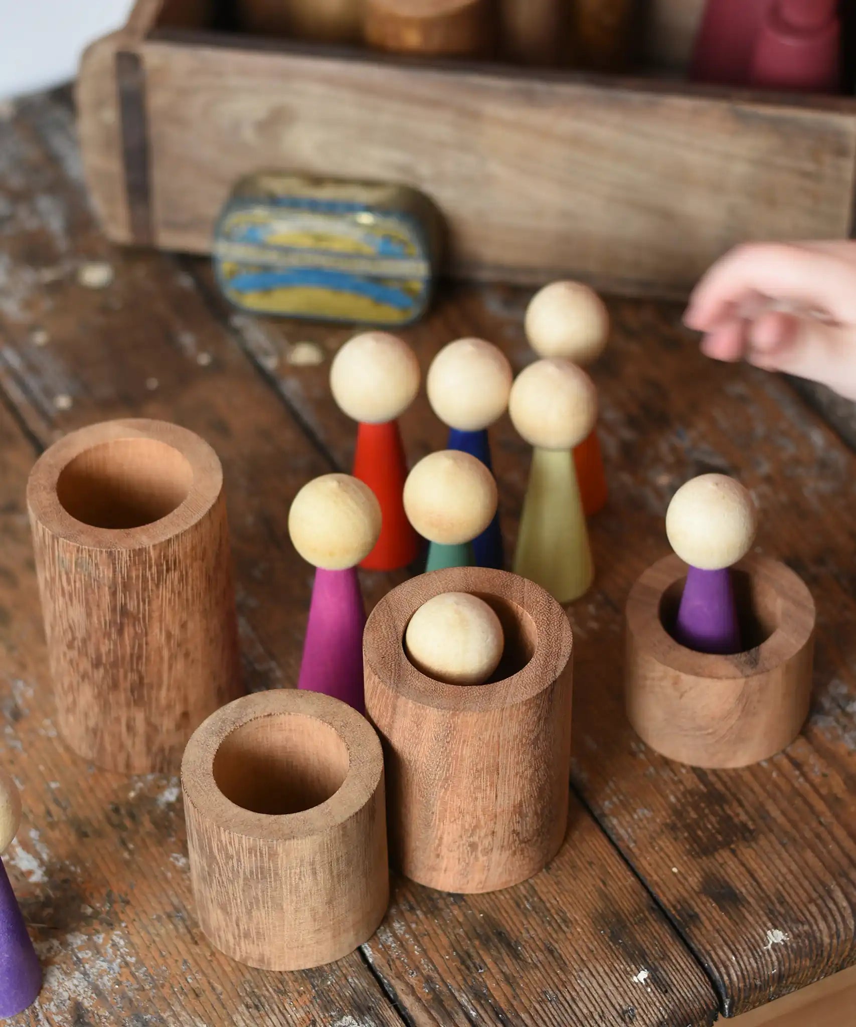 Papoose Toys 12 wooden Goethe rainbow people peg dolls placed on a wooden desk with some placed into wooden stacking tubes.