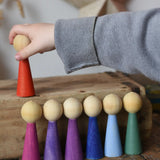 Papoose Toys 12 wooden Goethe rainbow people peg dolls placed on a wooden desk. Child's hand reaching for the red doll.