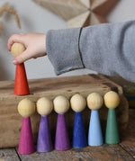 Papoose Toys 12 wooden Goethe rainbow people peg dolls placed on a wooden desk. Child's hand reaching for the red doll.