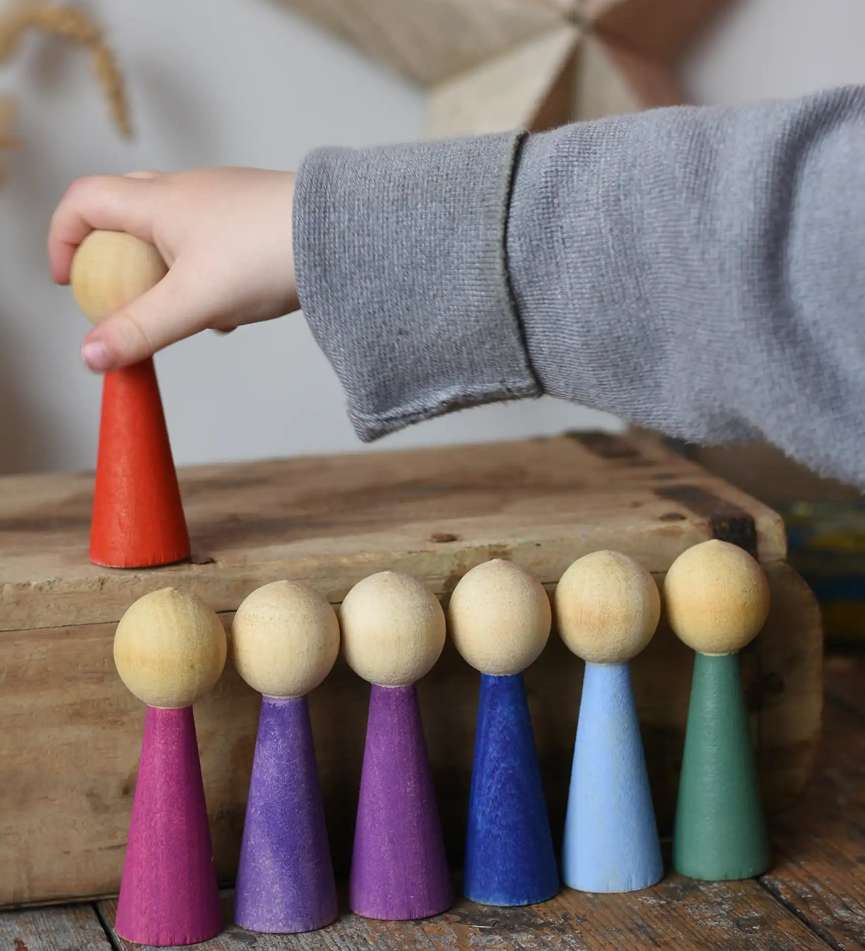 Papoose Toys 12 wooden Goethe rainbow people peg dolls placed on a wooden desk. Child's hand reaching for the red doll.