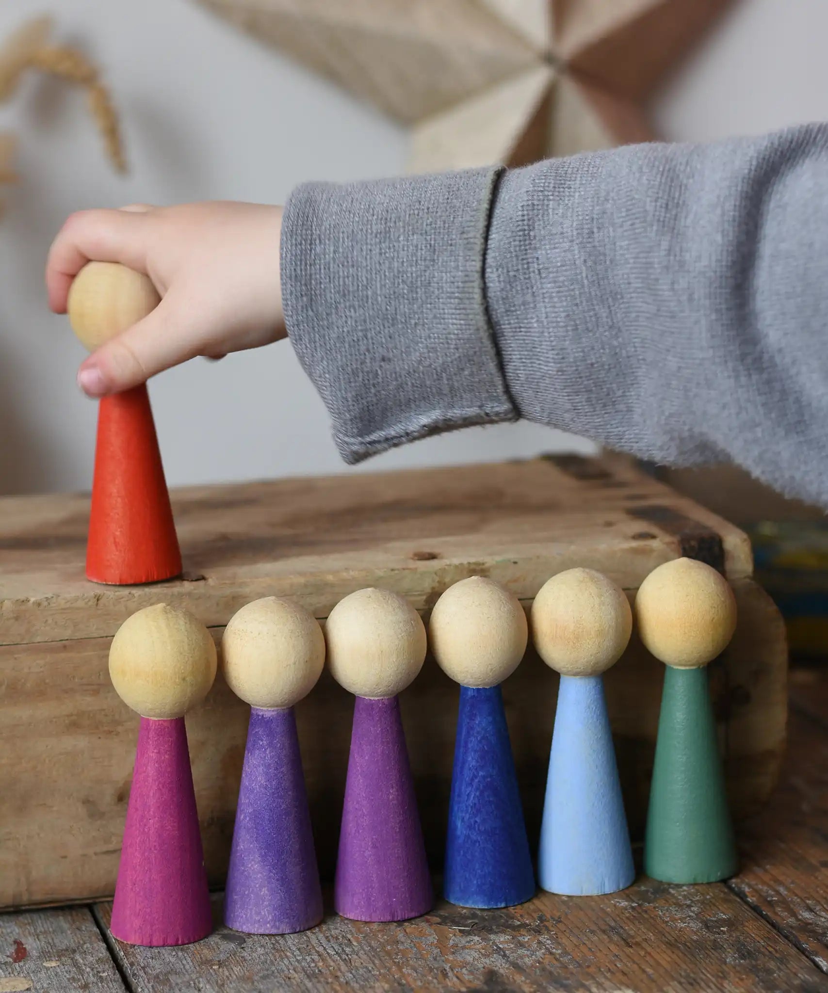 Papoose Toys 12 wooden Goethe rainbow people peg dolls placed on a wooden desk. Child's hand reaching for the red doll.