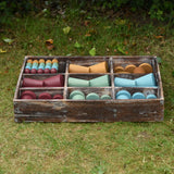 Papoose Toys wooden earth tinker tray school set placed on grass outdoors. The rustic look tray is filled with yellow, red, green and blue earth toned wooden toys and loose parts. 