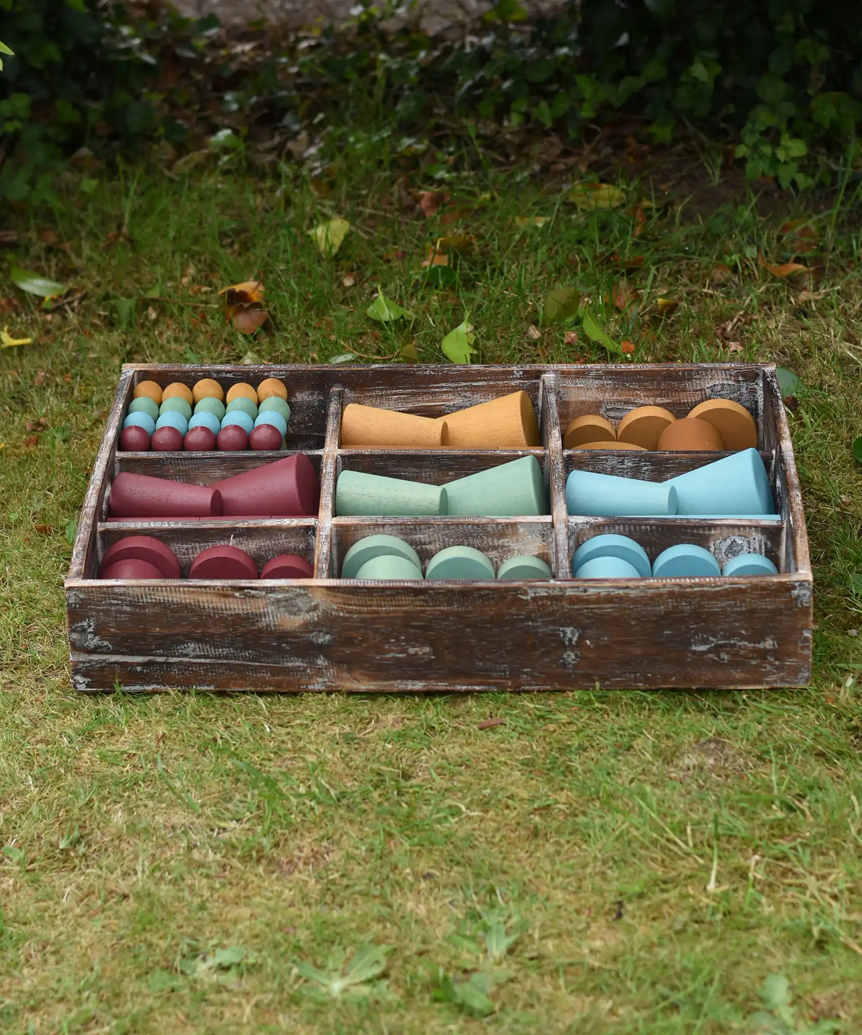 Papoose Toys wooden earth tinker tray school set placed on grass outdoors. The rustic look tray is filled with yellow, red, green and blue earth toned wooden toys and loose parts. 