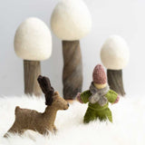 Papoose handmade felt reindeer figure and doll toy stood on a white fluffy blanket in front of 3 white Papoose trees