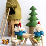 Two Papoose Toys Bearded Felt Tomten Gnome's sat at a wooden table and chairs with a felt Gnome house and pine tree in the background