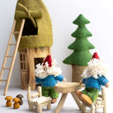 Two Papoose Toys Bearded Felt Tomten Gnome's sat at a wooden table and chairs with a felt Gnome house and pine tree in the background