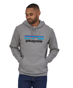 A man wearing a Patagonia Adult P-6 Logo Uprisal Hoody in a Gravel Heather grey colour stood in front of a plain background. 
