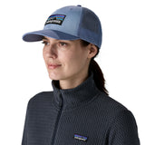 A woman wearing the Patagonia adult barnacle blue hat. A trucker style cap with a low profile in blue and features a navy mesh back with a logo embroidery on the front panel available at Babipur.