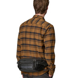 A man facing forwarding and wearing  the Patagonia waist pack in black around the waist . A 100% recycled material pack with 2 main compartments, side mesh pockets and a adjustable strap 