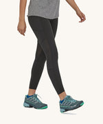 A woman walking in the black Patagonia running 7/8 tights on a cream background.