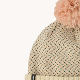 A close up of the Patagonia kids powder town beanie in confetti/natural. A thick beanie with blue and yellow speckled on natural coloured organic fabric. Features a light pink pom pom on top.