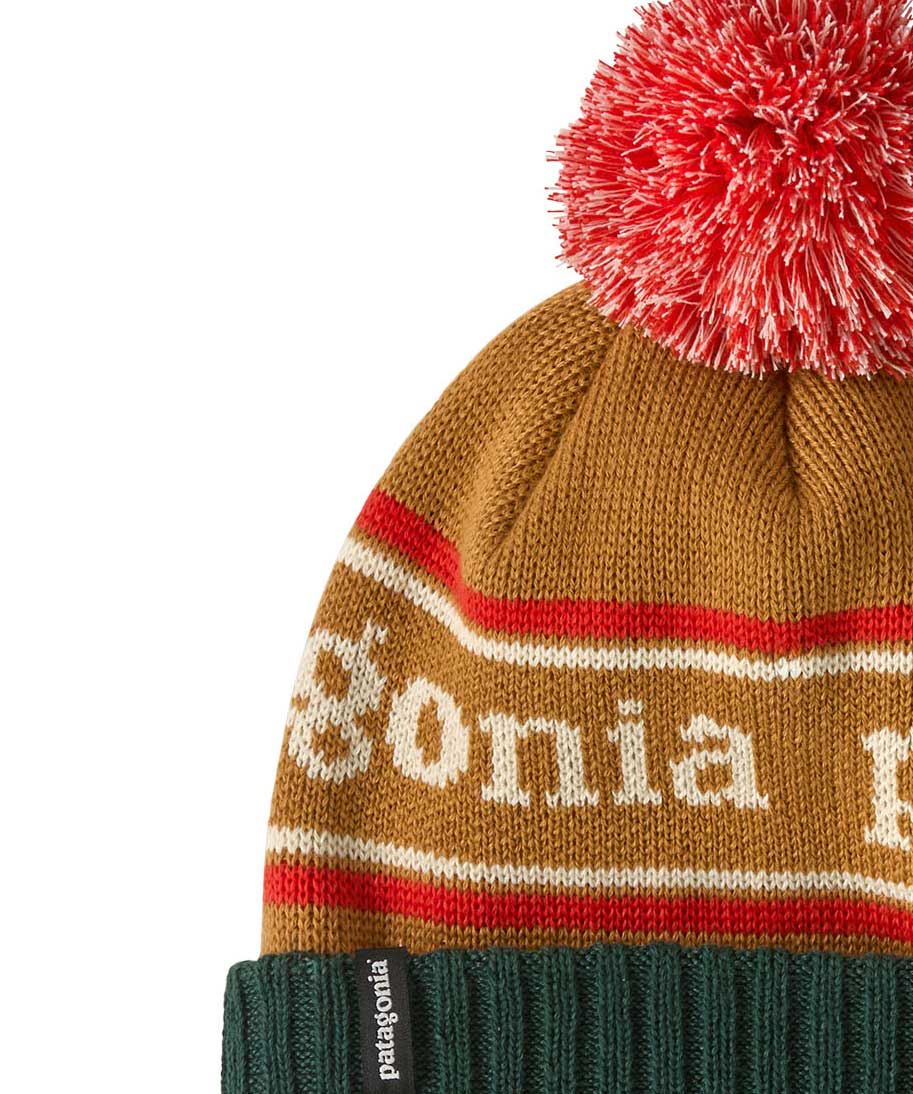 A close up of the Patagonia Kids  Powder Town Beanie in park stripe talon gold colour. A gold beanie with a green turn up brim. Features a red and white pompom on top and the brand name across the middle in white