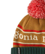 A close up of the Patagonia Kids  Powder Town Beanie in park stripe talon gold colour. A gold beanie with a green turn up brim. Features a red and white pompom on top and the brand name across the middle in white