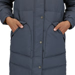 A close up of a person wearing the Patagonia Women's Downdrift Parka in Pine Smolder Blue. The image shows both hand in the side pockets and the zip and buttons fully closed