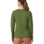 A woman wearing a Patagonia Women's long sleeved Cap Cool Daily Graphic Shirt in a Green X-Dye colour. The woman is stood in front of a plain background. The woman is facing away from the camera, showing the back of the top. 
