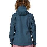 A woman wearing a Patagonia Women's Torrentshell 3L Rain Jacket in a Lagom Blue colour stood in front of a plain background. The woman is facing away showing the back of the coat. 