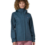 A woman wearing a Patagonia Women's Torrentshell 3L Rain Jacket in a Lagom Blue colour stood in front of a plain background. 