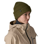 A woman wearing the Patagonia snowdrifter beanie in tent green with rolled up brim. Features a brand logo patch stitched on the side available at Babipur.