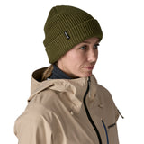 A woman wearing the Patagonia snowdrifter beanie in tent green with rolled up brim. Features a brand logo patch stitched on the side available at Babipur.