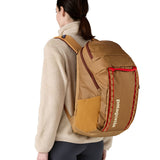 A woman wearing the Patagonia talon gold  32L black hole backpack. A 100% recycled polyester backpack in dark yellow with maroon and red detail. 