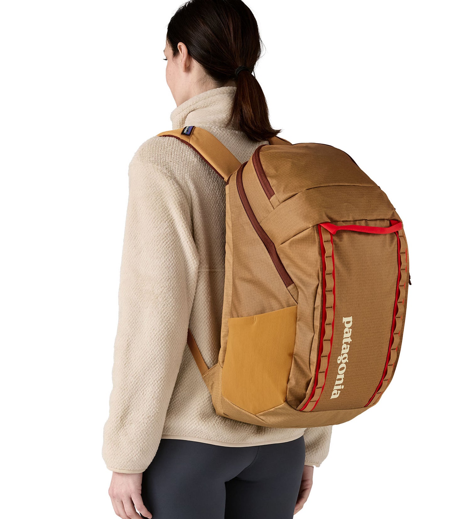 A woman wearing the Patagonia talon gold  32L black hole backpack. A 100% recycled polyester backpack in dark yellow with maroon and red detail. 