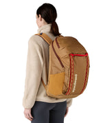 A woman wearing the Patagonia talon gold  32L black hole backpack. A 100% recycled polyester backpack in dark yellow with maroon and red detail. 