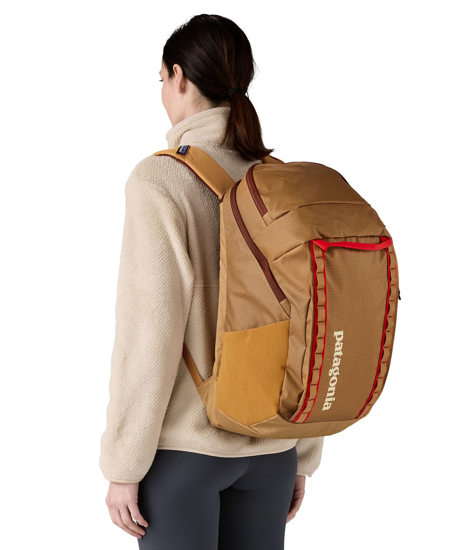 A woman wearing the Patagonia talon gold  32L black hole backpack. A 100% recycled polyester backpack in dark yellow with maroon and red detail. 